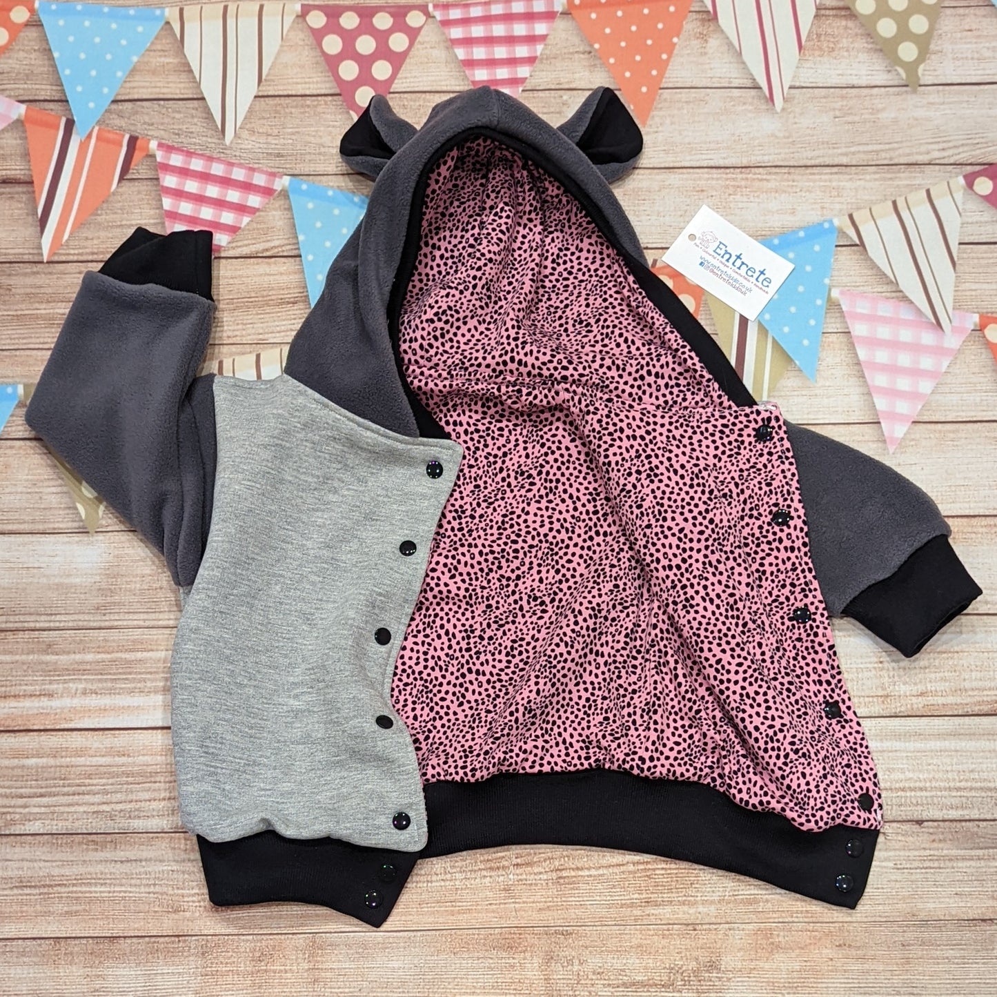 The reversible cat hoodie featuring fun tail and ear detailing. Handmade using grey polar fleece, light grey cotton french terry, black organic cotton jersey and graphite ribbing. The reverse is made using pink cheetah print cotton jersey. This cat hoodie is so unique and is great for imaginative play. Shown with the front popper entry open, revealing the pink cheetah reverse.