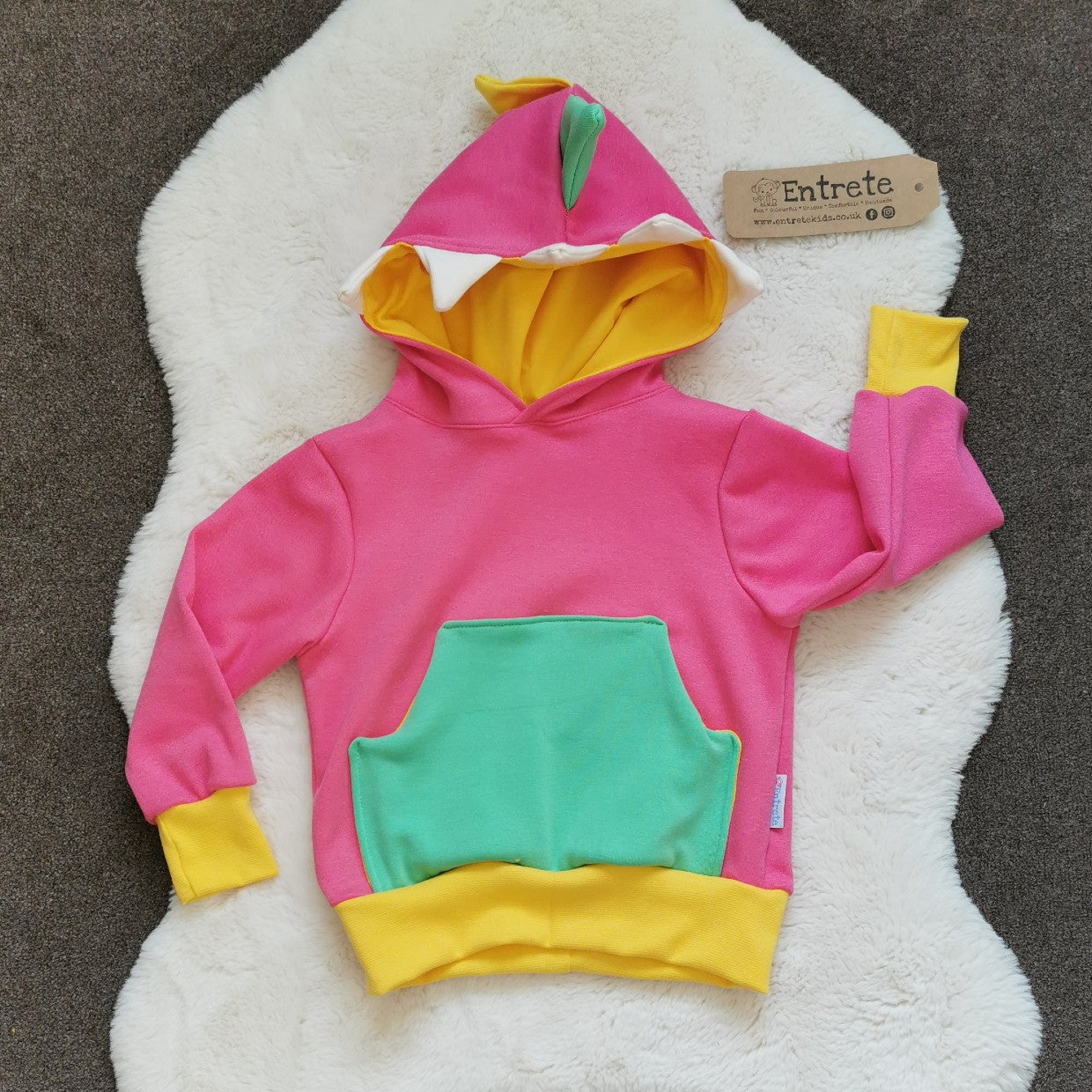 The pink dinosaur hoodie is packed full of colour and fun. Featuring a contrasting front pocket and dinosaur teeth and spikes on the hood. Handmade using pink, white and green cotton sweatshirt fleeces, with yellow cotton jersey and ribbing.