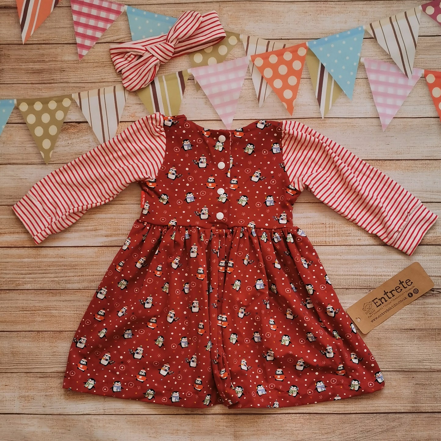 Rear view of a contrasting girls Christmas dress. Handmade using festive red penguins cotton jersey with contrasting red striped cotton jersey arms. Shown with a red striped headband (sold separately).