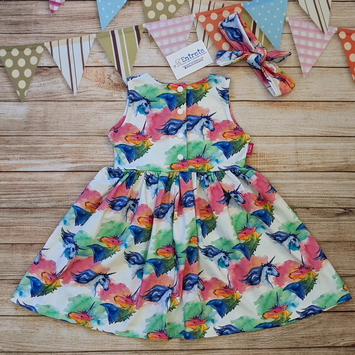 The gorgeously colourful unicorns on white sleeveless girls dress. Handmade using unicorns on white cotton jersey. Shown from the rear with a matching headband. The poppers run down to the waist as found in sizes over 9-12 months. Below 9-12 months the poppers run the length of the dress.