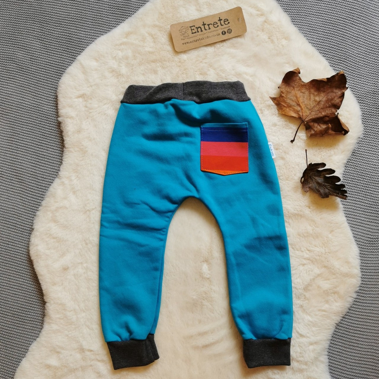 Rear view of harem joggers with elasticated waist, dropped crotch and roll-able ankle cuffs. Handmade using warm and vibrant sky blue cotton sweatshirt fleece, graphite ribbing and rainbow striped cotton jersey for the pocket.