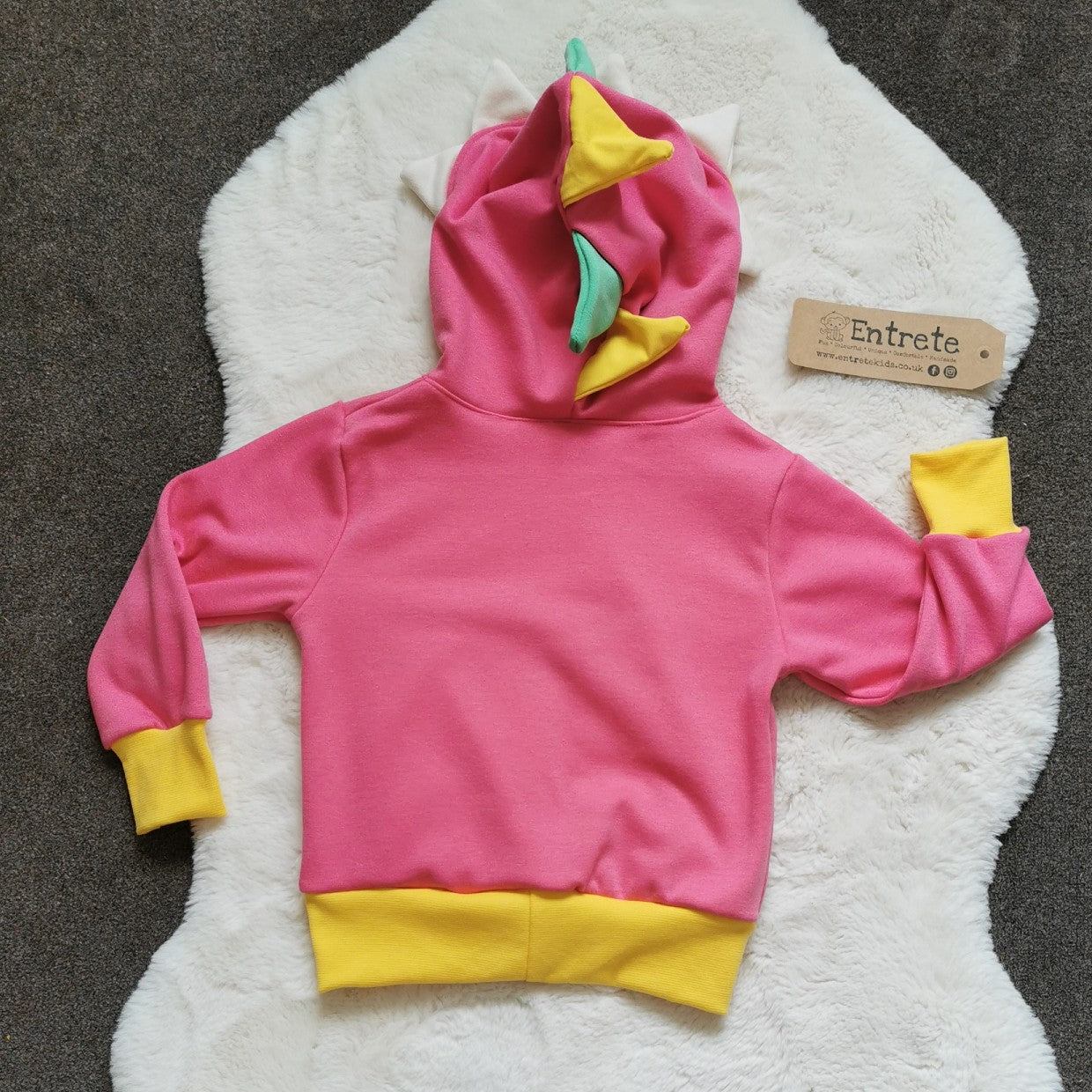 The pink dinosaur hoodie is packed full of colour and fun. Featuring a contrasting front pocket and dinosaur teeth and spikes on the hood. Handmade using pink, white and green cotton sweatshirt fleeces, with yellow cotton jersey and ribbing. Shown from the rear.