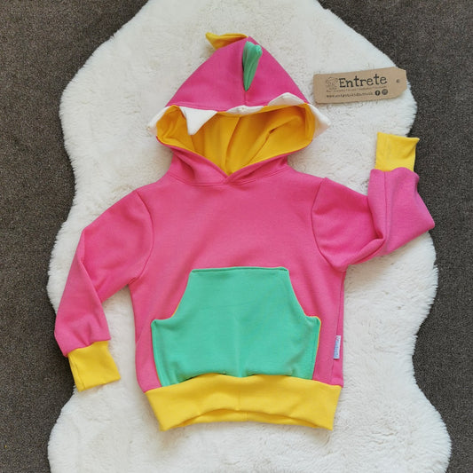 The pink dinosaur hoodie is packed full of colour and fun. Featuring a contrasting front pocket and dinosaur teeth and spikes on the hood. Handmade using pink, white and green cotton sweatshirt fleeces, with yellow cotton jersey and ribbing.