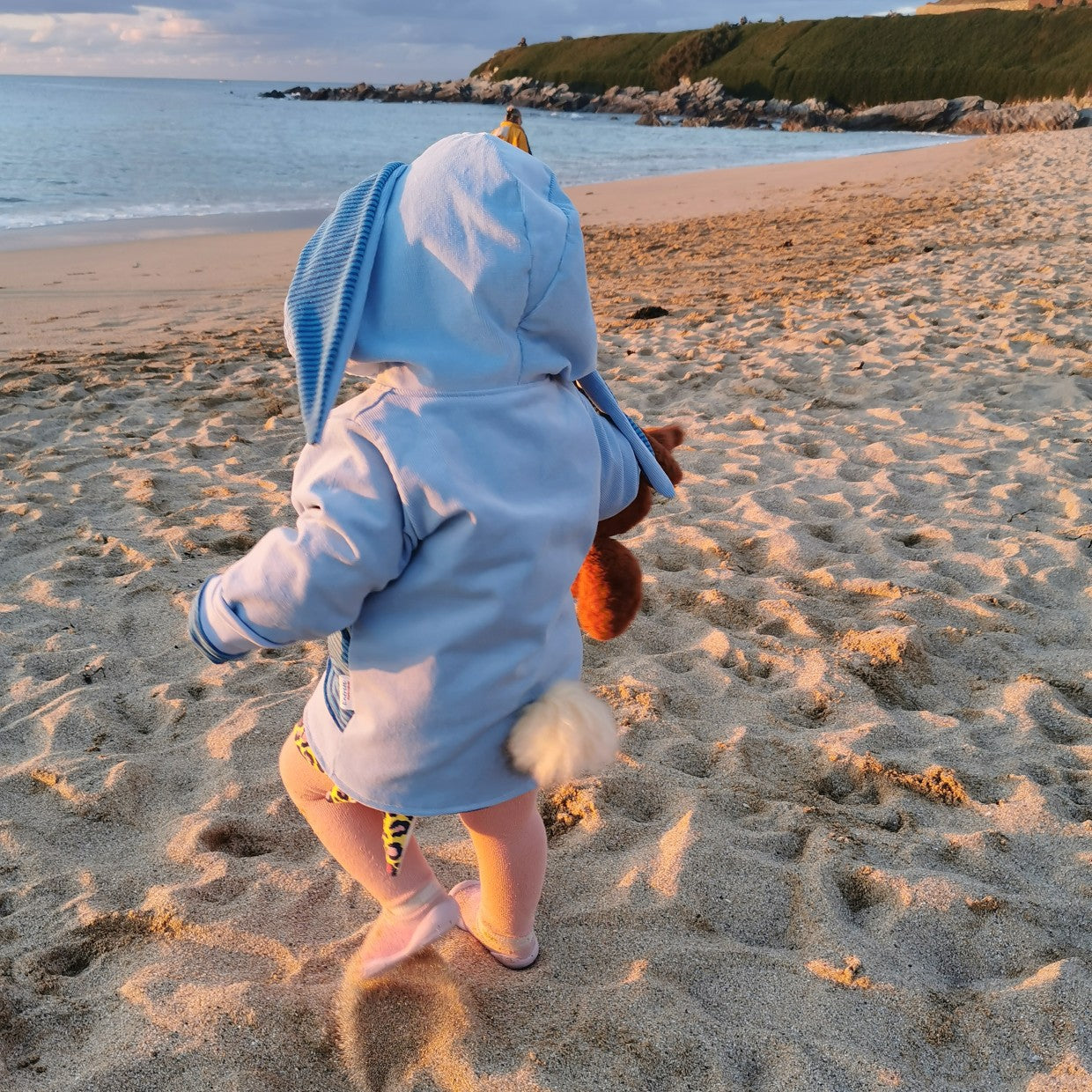 View of toddler wearing blue bunny coat on a beach. Showing bunny tail and ears.